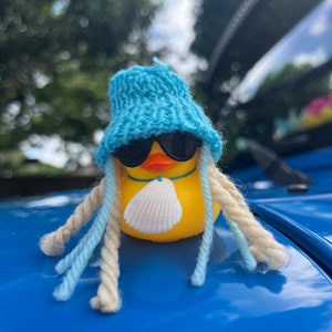 May include: A yellow rubber duck wearing a blue knitted hat, sunglasses, and a seashell necklace. The duck is sitting on a blue surface.