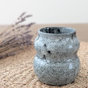 May include: A grey ceramic vase with a textured surface and a bulbous shape. The vase has a speckled pattern with dark grey and white accents. The interior is dark grey. The vase is on a woven mat.