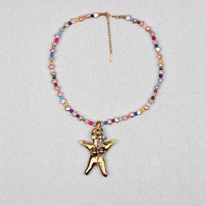 K&ouml;nnte beinhalten: Eine goldfarbene Halskette mit einem Anh&auml;nger in Form einer Person. Die Halskette hat eine Kette aus bunten Perlen. Der Anh&auml;nger hat ein detailliertes Design. Die Halskette wird vor einem hellgrauen Hintergrund gezeigt.