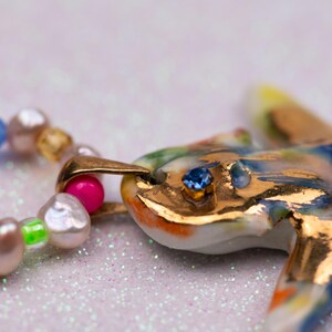 K&ouml;nnte beinhalten: Eine farbenfrohe, handgefertigte Halskette mit einem Keramik-Seestern-Anh&auml;nger mit goldenen Akzenten und einem blauen Edelsteinauge. Die Halskette enth&auml;lt eine Kette aus bunten Perlen, wodurch ein einzigartiges und k&uuml;nstlerisches Schmuckst&uuml;ck entsteht.