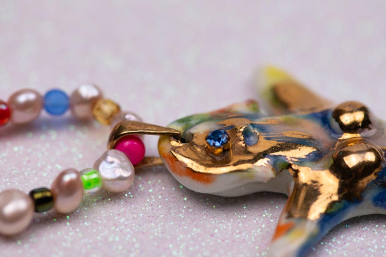 K&ouml;nnte beinhalten: Eine farbenfrohe, handgefertigte Halskette mit einem Keramik-Seestern-Anh&auml;nger mit goldenen Akzenten und einem blauen Edelsteinauge. Die Halskette enth&auml;lt eine Kette aus bunten Perlen, wodurch ein einzigartiges und k&uuml;nstlerisches Schmuckst&uuml;ck entsteht.