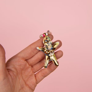 K&ouml;nnte beinhalten: Goldfarbene, abstrakte Figur in menschen&auml;hnlicher Form, gehalten in einer Hand vor rosa Hintergrund. Die Figur hat blaue, gelbe und rosa Akzente und ist mit einer Perlenkette verziert. Das Gesamtdesign ist k&uuml;nstlerisch und einzigartig.