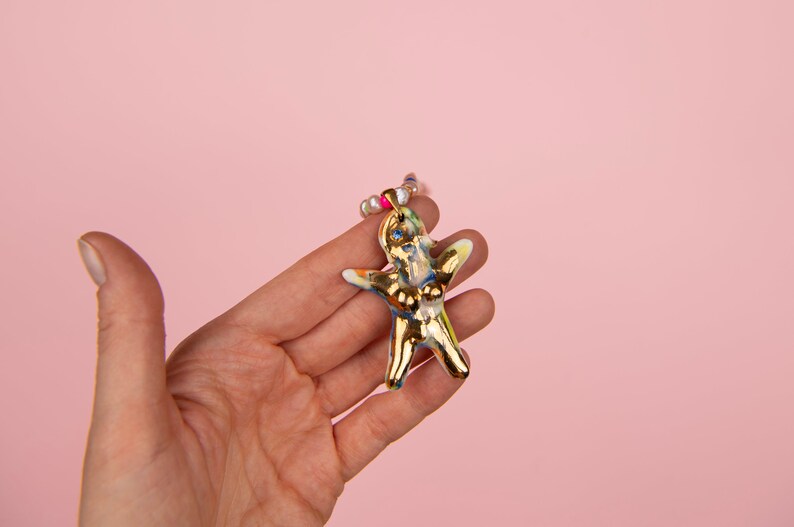 K&ouml;nnte beinhalten: Goldfarbene, abstrakte Figur in menschen&auml;hnlicher Form, gehalten in einer Hand vor rosa Hintergrund. Die Figur hat blaue, gelbe und rosa Akzente und ist mit einer Perlenkette verziert. Das Gesamtdesign ist k&uuml;nstlerisch und einzigartig.