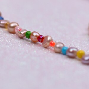 K&ouml;nnte beinhalten: Nahaufnahme einer Halskette mit rosa Perlen und bunten Perlen. Die Perlen sind in verschiedenen Farben gehalten, darunter Rosa, Gr&uuml;n, Blau, Gelb und Lila. Die Halskette liegt auf einem hellrosa, glitzernden Hintergrund.