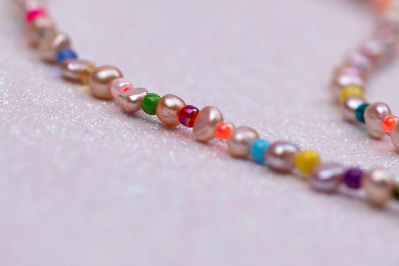 K&ouml;nnte beinhalten: Nahaufnahme einer Halskette mit rosa Perlen und bunten Perlen. Die Perlen sind in verschiedenen Farben gehalten, darunter Rosa, Gr&uuml;n, Blau, Gelb und Lila. Die Halskette liegt auf einem hellrosa, glitzernden Hintergrund.