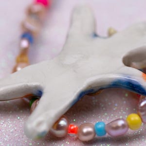 K&ouml;nnte beinhalten: Handgefertigte Halskette mit einem wei&szlig;en Keramik-Seestern-Anh&auml;nger mit blauen und orangen Akzenten. Die Halskette enth&auml;lt eine Kette aus bunten Perlen und S&uuml;&szlig;wasserperlen, wodurch ein einzigartiges Schmuckst&uuml;ck entsteht.
