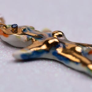 K&ouml;nnte beinhalten: Nahaufnahme eines handgefertigten Keramik-Anh&auml;ngers mit goldenen Akzenten und einem kleinen blauen Edelstein. Der Anh&auml;nger ist wei&szlig; mit blauen und orangefarbenen Details. Die Halskette hat auch rosa und wei&szlig;e Perlen.