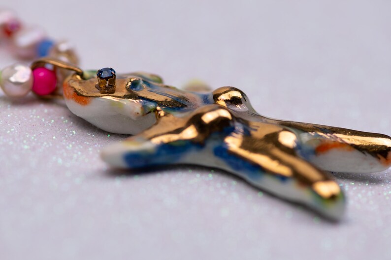 K&ouml;nnte beinhalten: Nahaufnahme eines handgefertigten Keramik-Anh&auml;ngers mit goldenen Akzenten und einem kleinen blauen Edelstein. Der Anh&auml;nger ist wei&szlig; mit blauen und orangefarbenen Details. Die Halskette hat auch rosa und wei&szlig;e Perlen.