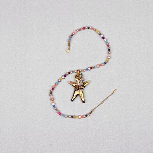 K&ouml;nnte beinhalten: Eine goldfarbene Anh&auml;ngerkette mit einem stilisierten menschlichen Figurenanh&auml;nger. Der Anh&auml;nger h&auml;ngt an einer Kette aus bunten, unregelm&auml;&szlig;ig geformten Perlen. Die Kette wird auf einer hellgrauen Oberfl&auml;che pr&auml;sentiert.