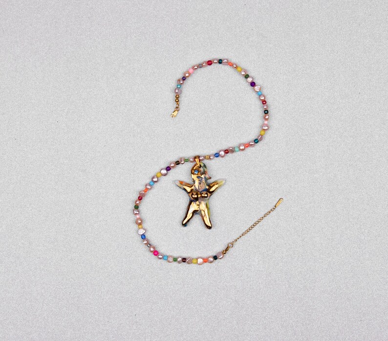 K&ouml;nnte beinhalten: Eine goldfarbene Anh&auml;ngerkette mit einem stilisierten menschlichen Figurenanh&auml;nger. Der Anh&auml;nger h&auml;ngt an einer Kette aus bunten, unregelm&auml;&szlig;ig geformten Perlen. Die Kette wird auf einer hellgrauen Oberfl&auml;che pr&auml;sentiert.