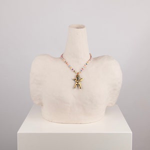 K&ouml;nnte beinhalten: Eine goldfarbene Anh&auml;ngerkette mit einem figurf&ouml;rmigen Anh&auml;nger wird auf einer wei&szlig;en B&uuml;ste pr&auml;sentiert. Die Kette hat eine Perlenkette mit bunten Perlen. Die B&uuml;ste steht auf einem wei&szlig;en Sockel.