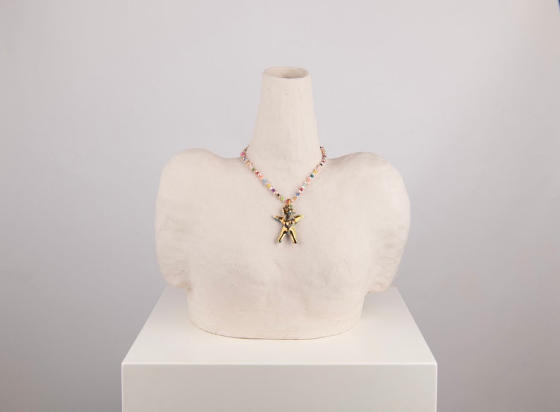 K&ouml;nnte beinhalten: Eine goldfarbene Anh&auml;ngerkette mit einem figurf&ouml;rmigen Anh&auml;nger wird auf einer wei&szlig;en B&uuml;ste pr&auml;sentiert. Die Kette hat eine Perlenkette mit bunten Perlen. Die B&uuml;ste steht auf einem wei&szlig;en Sockel.