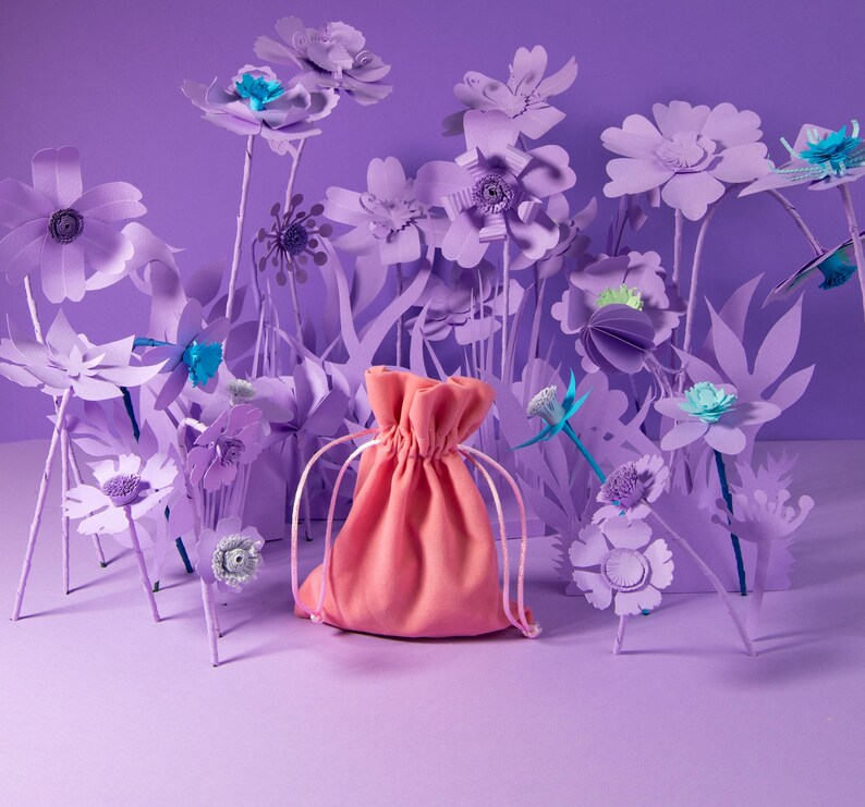 K&ouml;nnte beinhalten: Ein korallenfarbener Kordelzugbeutel steht vor einem Hintergrund aus Papierblumen in verschiedenen Lilat&ouml;nen. Die Blumen haben t&uuml;rkisfarbene und wei&szlig;e Akzente und schaffen eine skurrile, k&uuml;nstlerische Szene. Der Beutel ist der Mittelpunkt.
