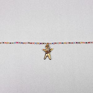 K&ouml;nnte beinhalten: Eine goldfarbene Anh&auml;ngerkette mit einer stilisierten menschlichen Figur. Der Anh&auml;nger h&auml;ngt an einer zarten Kette aus kleinen, bunten Perlen. Die Halskette wird vor einem hellgrauen Hintergrund pr&auml;sentiert.