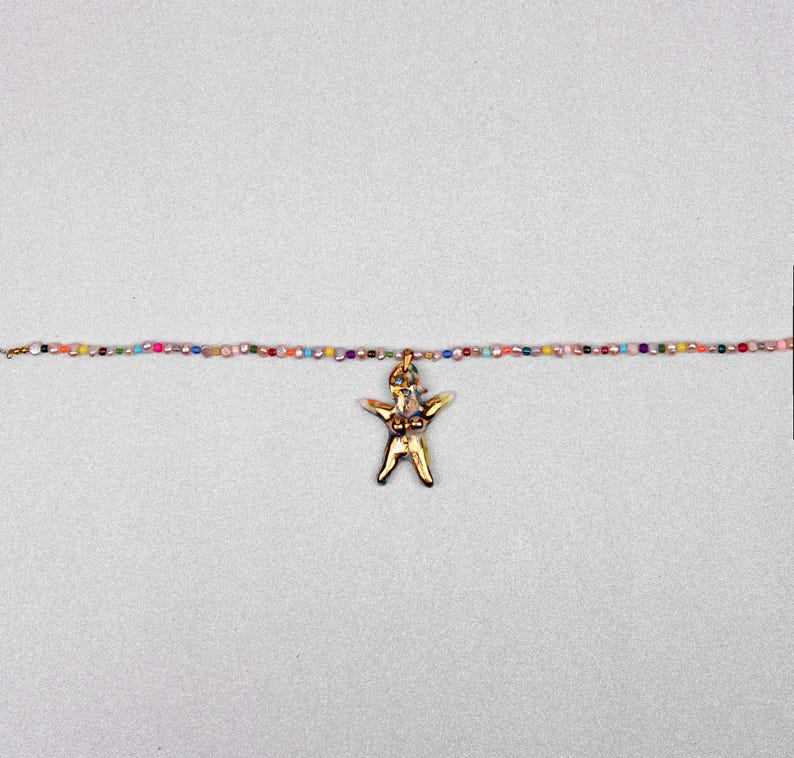 K&ouml;nnte beinhalten: Eine goldfarbene Anh&auml;ngerkette mit einer stilisierten menschlichen Figur. Der Anh&auml;nger h&auml;ngt an einer zarten Kette aus kleinen, bunten Perlen. Die Halskette wird vor einem hellgrauen Hintergrund pr&auml;sentiert.