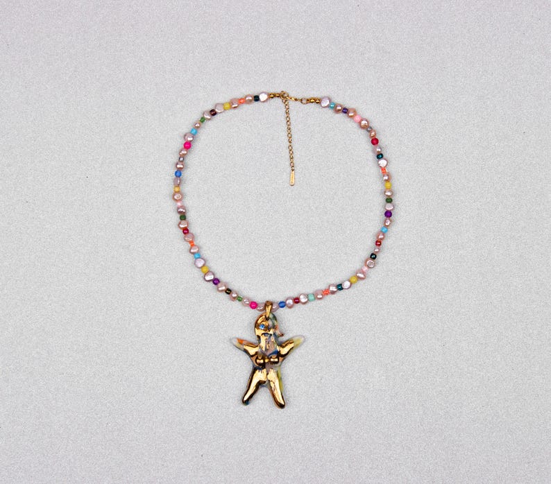 K&ouml;nnte beinhalten: Eine goldfarbene Halskette mit einem Anh&auml;nger in Form einer Person. Die Halskette hat eine Kette aus bunten Perlen. Der Anh&auml;nger hat ein detailliertes Design. Die Halskette wird vor einem hellgrauen Hintergrund gezeigt.