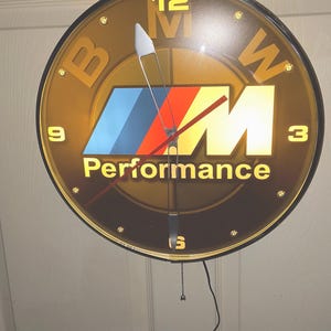 May include: A round, illuminated wall clock featuring the BMW M Performance logo in blue, red, and yellow. The clock face has a dark amber background with yellow numerals at 12, 3, 6, and 9 o'clock, silver hands, a red second hand, and a pull chain.
