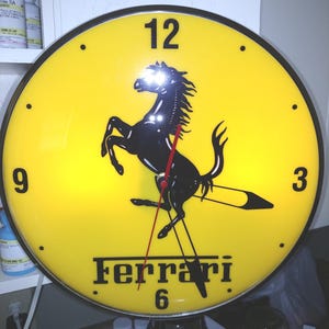 May include: A round wall clock with a bright yellow face featuring a black prancing horse logo in the center. The clock has black numbers at the 12, 3, 6, and 9 positions, the word Ferrari in black text, and thin red hour and minute hands.