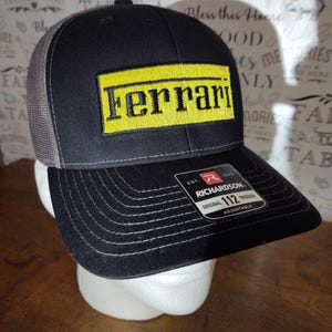 May include: A black and gray trucker hat with a yellow patch that says "Ferrari" on it. The hat is adjustable and has a Richardson logo on the back.