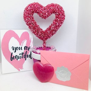 May include: Pink glitter heart shaped decoration with a pink envelope and a card that says "you are beautiful".