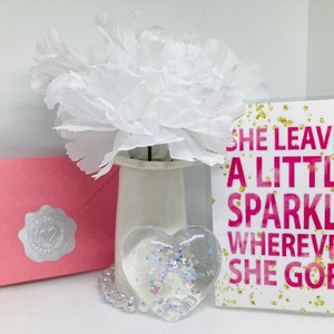 May include: A white card with gold glitter and pink text that reads "She leaves a little sparkle wherever she goes". A white flower, a pink envelope with a silver seal, and a clear heart-shaped resin ornament with iridescent glitter are also in the image.