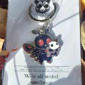 May include: A black and red moth keychain with a small white animal in its arms. The keychain is attached to a silver ring with a black and white raccoon charm. The keychain is on a white card with the text "We're all weird sometimes!" and the website address "www.mountainsmissfits.com".