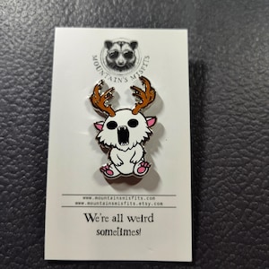 May include: White enamel pin with a cartoon white creature with pink paws and antlers. The creature has a wide open mouth and is wearing a silver collar. The pin is on a white card with the text "Mountain's Misfits" and "We're all weird sometimes!"