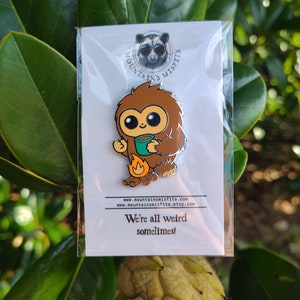May include: A brown and white enamel pin featuring a cartoon creature holding a green mug. The creature is sitting by a campfire. The pin says "We're all weird sometimes!" and includes the website address "www.mountainsmistsfits.com".
