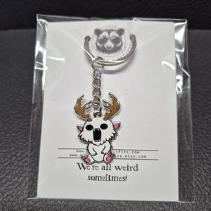 May include: A silver-toned metal keychain featuring a white creature with brown antlers, a wide-open mouth, and pink accents. The keychain is attached to a white display card with a raccoon graphic and the text "We're all weird sometimes!"