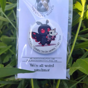 May include: A round button with a cartoon moth and opossum illustration, with the text "IT'S DANGEROUS TO GO ALONE. TAKE THIS!" and "We're all weird sometimes!". The button is packaged on a white card.