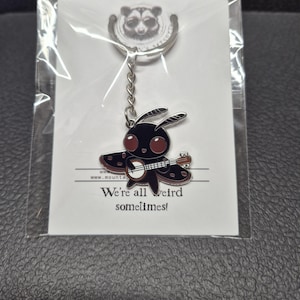 May include: A metal keychain featuring a black moth character with large dark red eyes playing a banjo. The keychain is attached to a white backing card that displays a raccoon logo and the text "We're all weird sometimes!" in black ink.