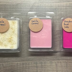 May include: Three wax melt packages in clear plastic clamshells. The first contains a cream-colored wax with gold glitter and a label that reads "Vanilla Caramel." The second is pink with the same label. The third is hot pink with a label that reads "Strawberry Pound Cake."