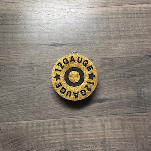 May include: A gold-colored, circular ornament with the text "12 GAUGE" in black lettering. The ornament has a black center and a small metal loop for hanging. The background is a wooden surface.