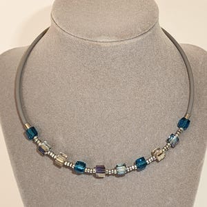 May include: A silver choker necklace with a blue and silver beaded design. The necklace is on a grey mannequin.