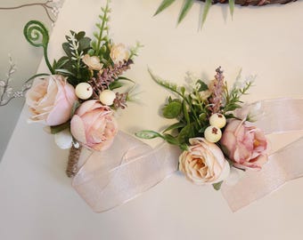 Könnte beinhalten: Zwei florale Ansteckblumen mit zartrosa künstlichen Rosen, kleinen weißen Beeren, Lavendelzweigen und grünem Blattwerk. Jedes Arrangement ist mit einem schimmernden, hellen Band und einem umwickelten Stielansatz zur Befestigung versehen.