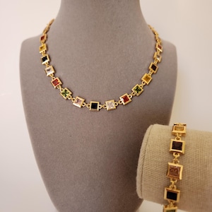 Pode incluir: Um conjunto de colar e pulseira dourados. As joias apresentam pedras preciosas quadradas multicoloridas em metal dourado. O colar é exibido em um manequim cinza e a pulseira em um tecido bege.