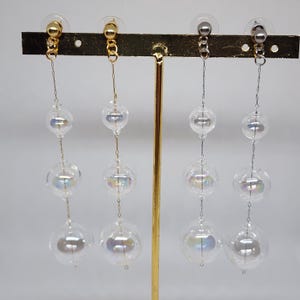 May include: Four pairs of dangle earrings with clear, iridescent glass orbs suspended from gold and silver-toned posts. The earrings are displayed on a gold-toned stand against a white background. Each earring features three orbs.