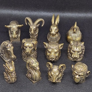 Puede incluir: Una colección de once pequeñas figuras de metal color latón que representan varias cabezas de animales, incluyendo un toro, carnero, conejo, gallo, caballo, perro, cerdo, tigre, dragón, serpiente, ratón y mono, con detalles grabados.