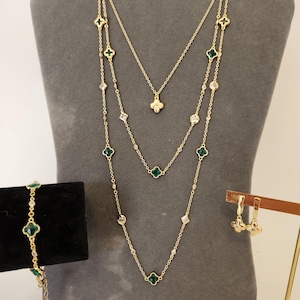 May include: A gold-toned jewelry set consisting of a multi-strand necklace, a bracelet, and a pair of drop earrings. Each piece features small green clover-shaped charms and clear, round, and diamond-shaped crystals set along delicate gold chains.