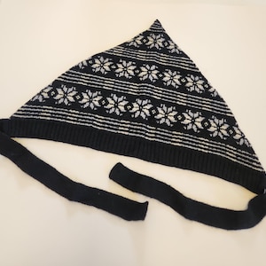 May include: A triangular knit headscarf in dark navy blue featuring a repeating white and light blue snowflake pattern. The garment has a ribbed bottom edge and two long, solid dark navy blue knit ties extending from the bottom corners.