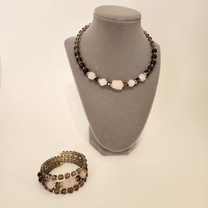 May include: A set of two necklaces featuring brown and white beads with a pink stone accent. The necklace is on a gray mannequin and the bracelet is on a white surface.