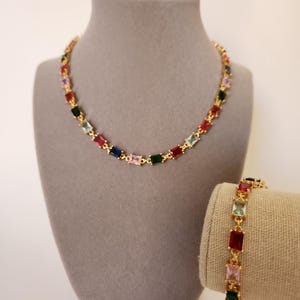 May include: A gold-toned necklace and bracelet set. The necklace is displayed on a gray bust, and the bracelet is on a tan fabric. Both pieces feature colorful, rectangular gemstones in various colors.