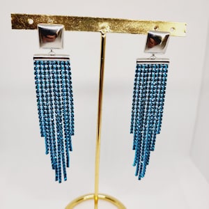 Puede incluir: Un par de pendientes largos de plata con flecos de cristal azul. Los pendientes se muestran en un soporte dorado.