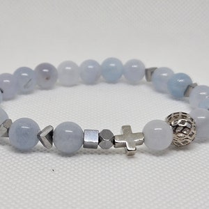Könnte beinhalten: Ein hellblaues Edelstein-Armband mit silbernen Akzenten, darunter ein Kreuzanhänger und eine dekorative Kugelperle.