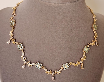 May include: A gold-toned necklace with a floral design. The necklace features small, light blue flowers, clear teardrop-shaped accents, and delicate leaf details. The necklace is displayed on a gray mannequin.