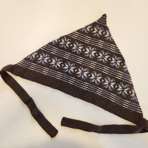 May include: A triangular knit headscarf in dark brown featuring a repeating pattern of light blue and white snowflakes. The base of the scarf has a ribbed texture and includes two long, solid dark brown knit ties for securing the garment.