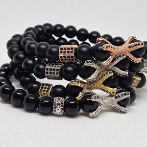 Könnte beinhalten: Vier schwarze Perlenarmbänder mit silbernen, goldenen und roségoldenen Akzenten. Jedes Armband hat einen einzigartigen Anhänger mit schwarzen Steinen.