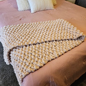 May include: A soft, blush pink throw blanket with a chunky knit texture. The blanket is draped over a bed with white and cream pillows.