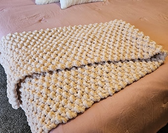 Può includere: Una morbida coperta rosa tenue con una trama a maglia grossa. La coperta è drappeggiata su un letto con cuscini bianchi e crema.