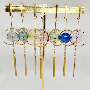 May include: Six pairs of gold hoop earrings with a dangling glass orb and a gold chain. The orbs are in various colors, including light blue, green, pink, yellow, blue, and purple.